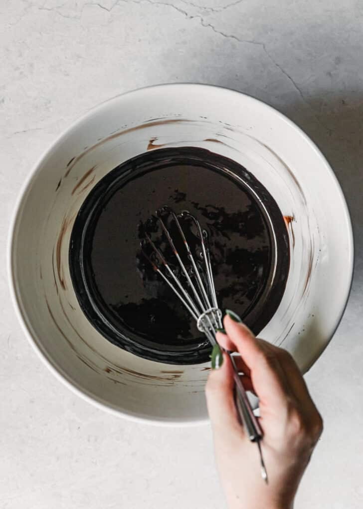 A hand whisking butter, chocolate, and cocoa powder in a white bowl on a white background.