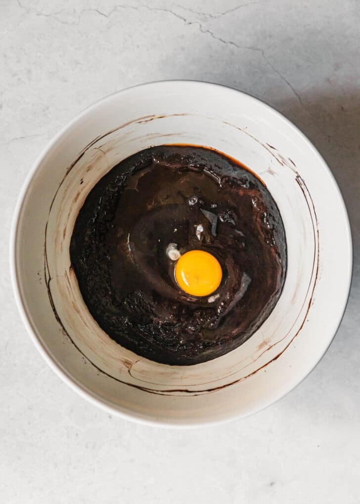 A white bowl with chocolate, oil, and eggs on a white table.