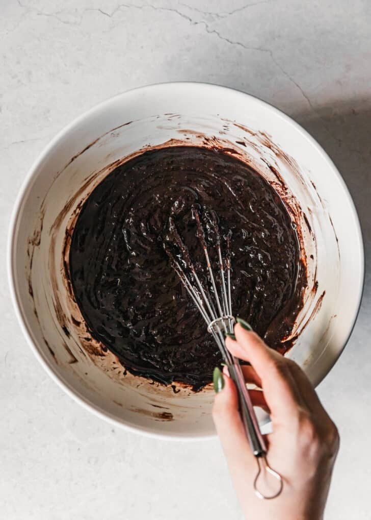 A hand whisking chocolate batter in a white bowl on a white counter.