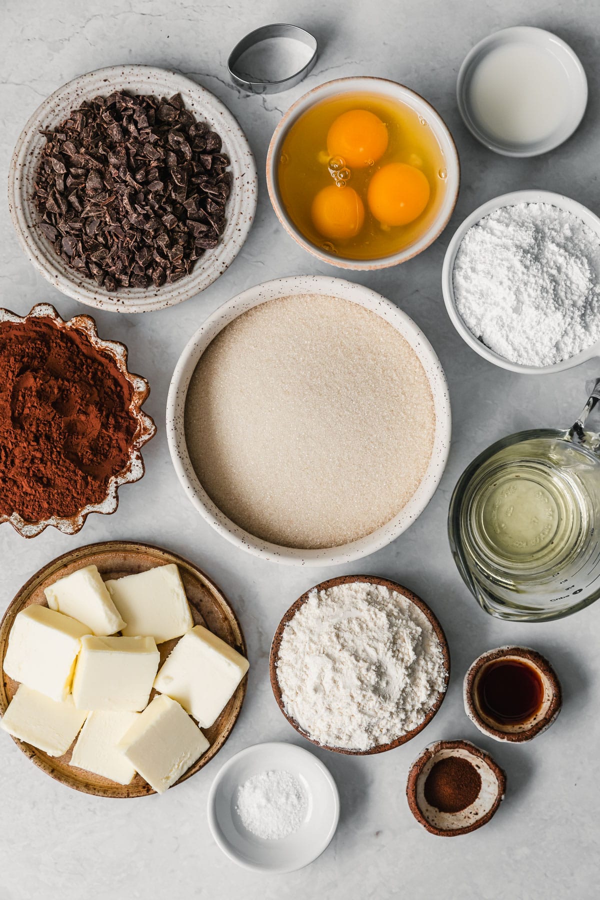 White and brown bowls of sugar, cocoa, powdered sugar, eggs, chocolate, flour, butter, oil, vanilla, and salt on a white counter.