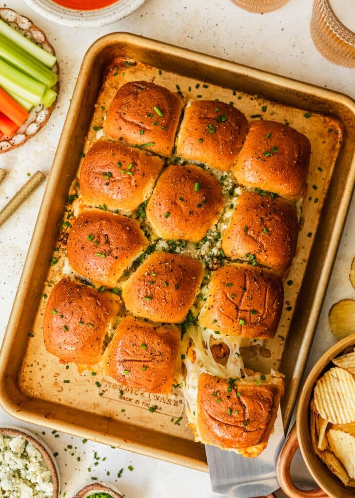 A spatula grabbing a slider from a tray of buffalo chicken sliders on a beige table next to glasses of beer, a brown bowl of chips, and a white plate of carrots and celery.