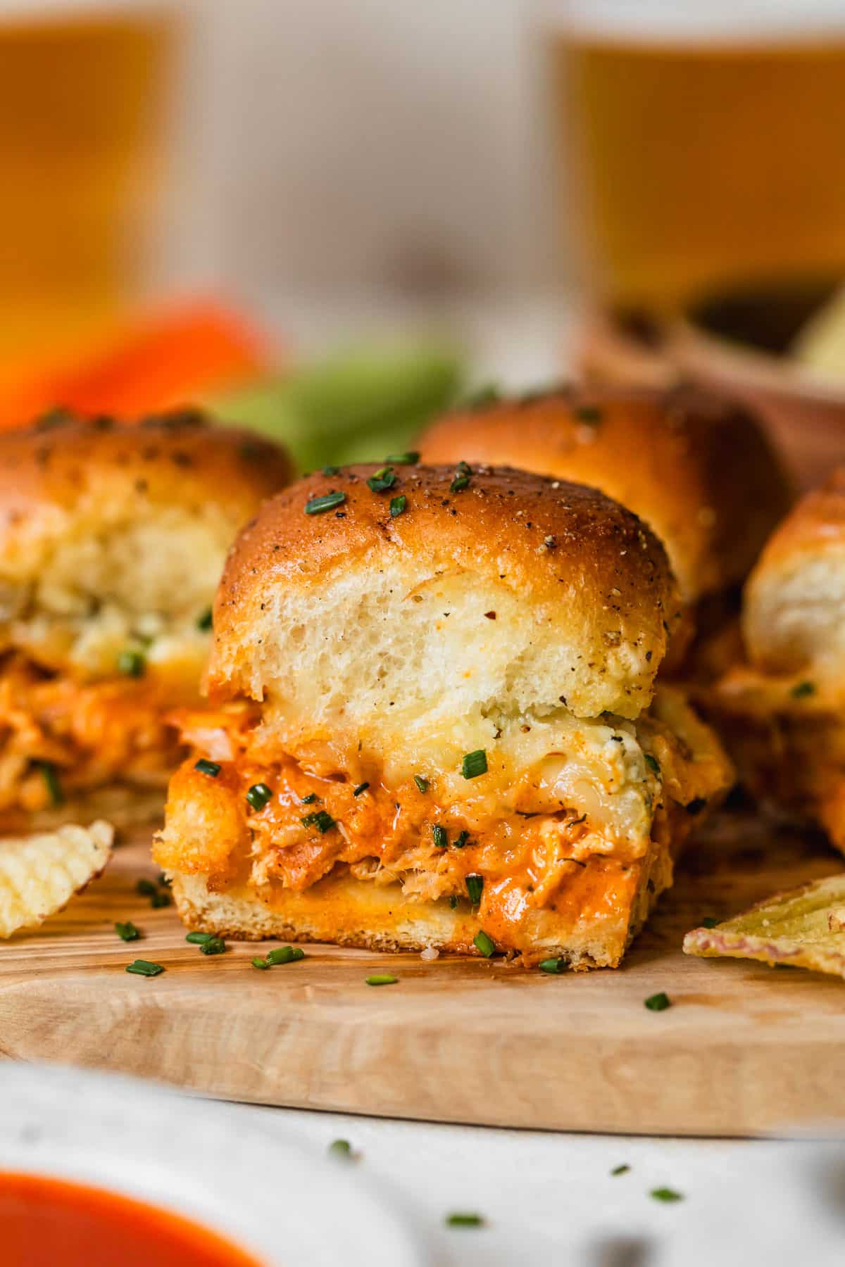 Buffalo chicken sliders on a wood board next to a white bowl of buffalo sauce and glasses of beer with a white background.