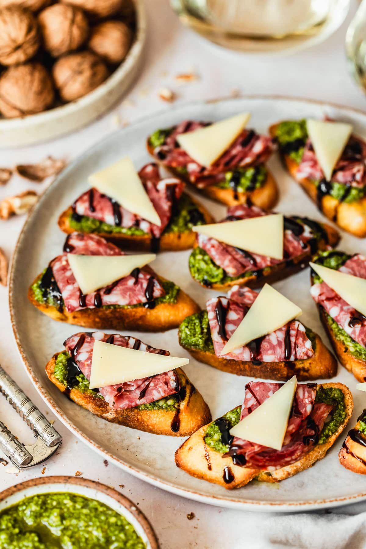 A grey platter of soppressata, parmesan, and pesto crostini on a tan counter next to glasses of white wine, a white bowl of pesto, and a white bowl of walnuts.