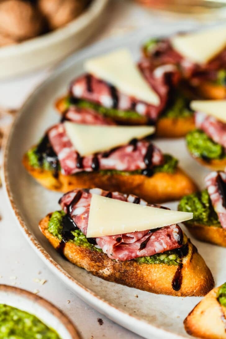 A grey platter of pesto crostini with salami and parmesan on a tan counter next to glasses of white wine and a white platter of walnuts.