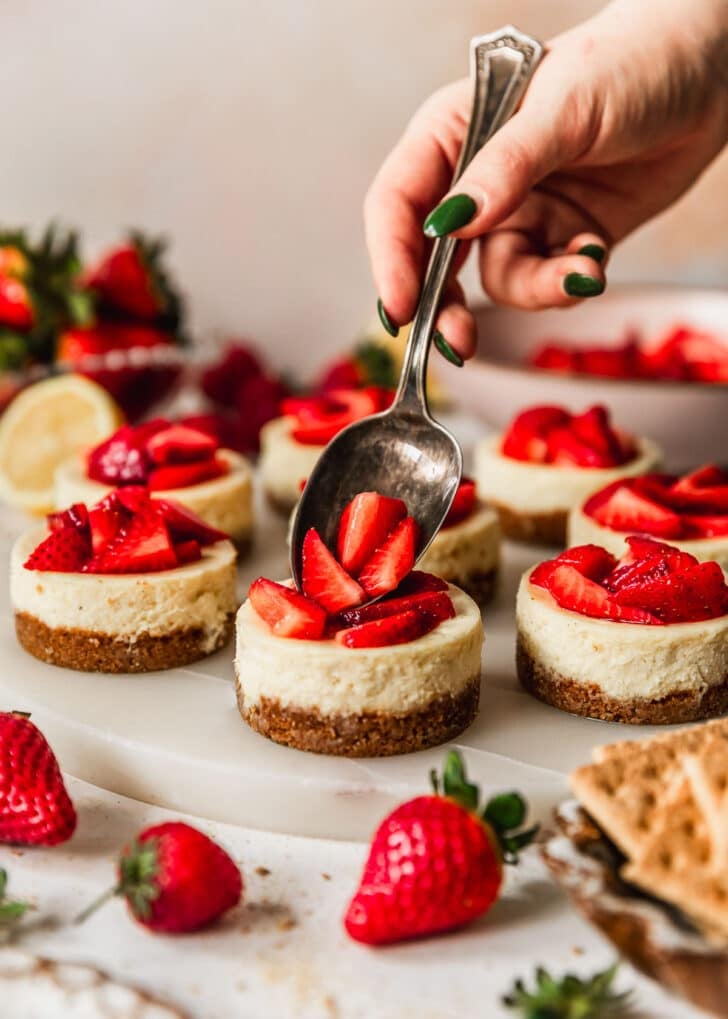 A hand spooning strawberry topping on mini cheesecakes next to a pink bowl, strawberries, and a brown bowl of graham crackers on a pink background.