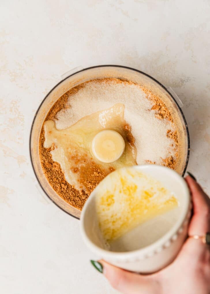 A hand pouring a bowl of melted butter into a food processor of graham crumbs.