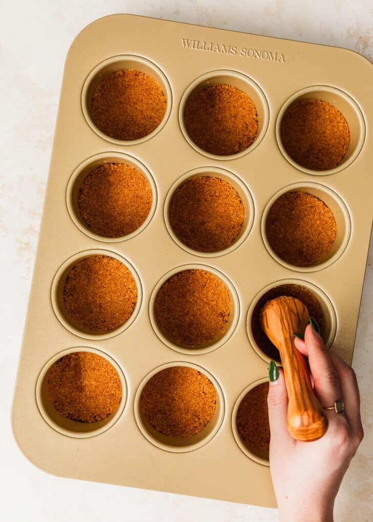 A hand using a pastry tamper to press graham cracker crust into a mini cheesecake pan.