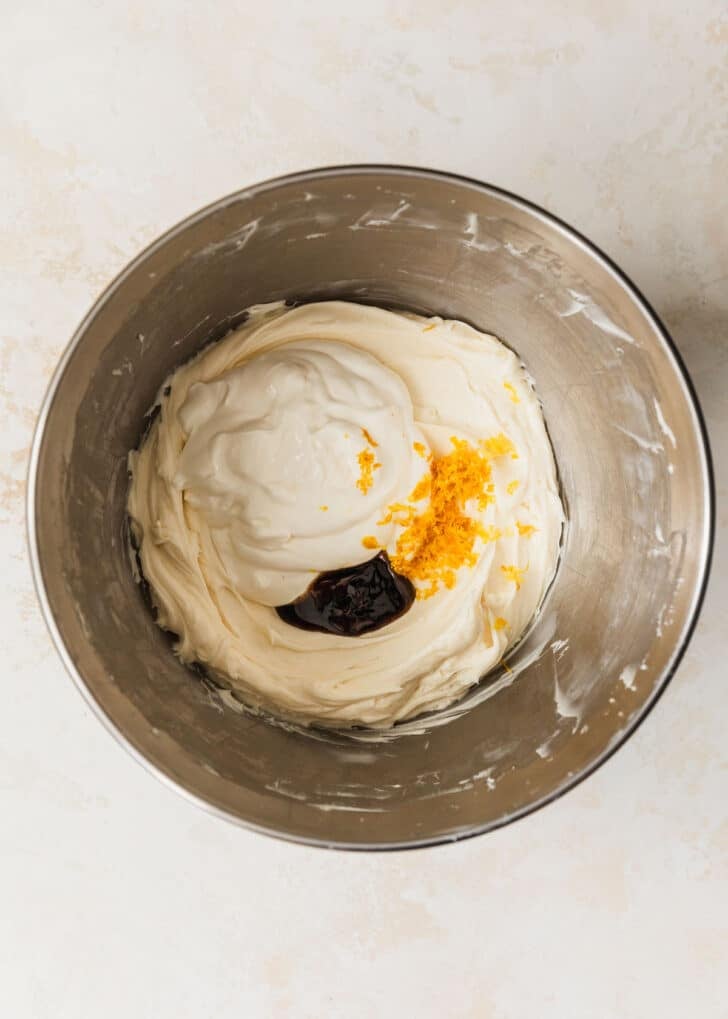 A silver bowl of cream cheese filling, lemon zest, sour cream, and vanilla on a tan counter.