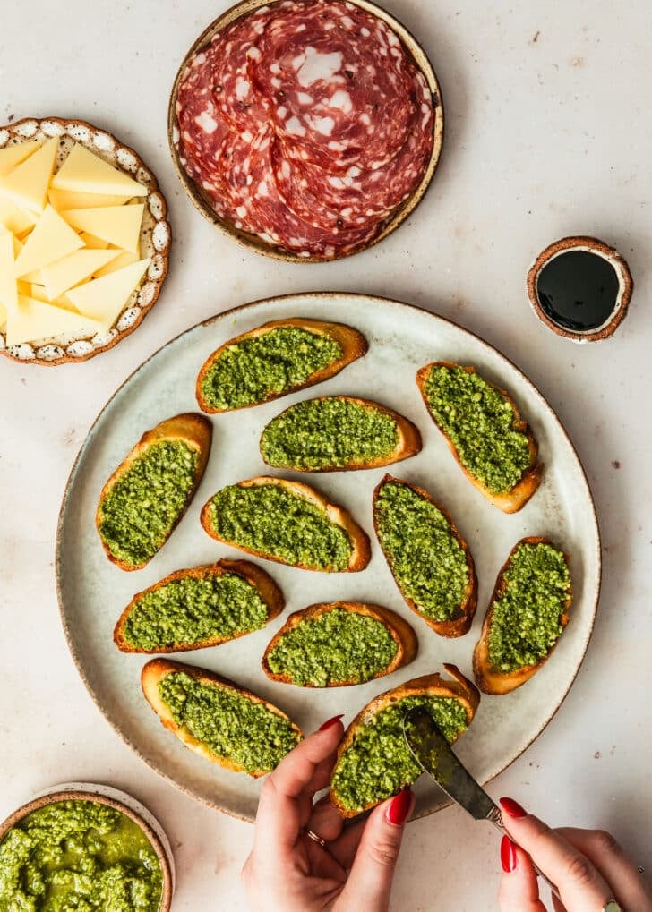 A hand spreading basil sauce on toasts on a grey plate next to brown bowls of soppressata, parmesan, and balsamic glaze with a tan background.