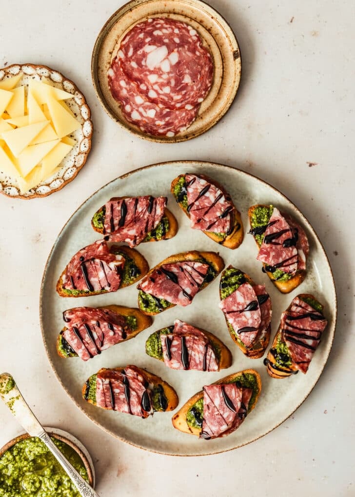 A grey platter of toasts with salami on a tan counter next to brown bowls of green sauce, parmesan, and salami.