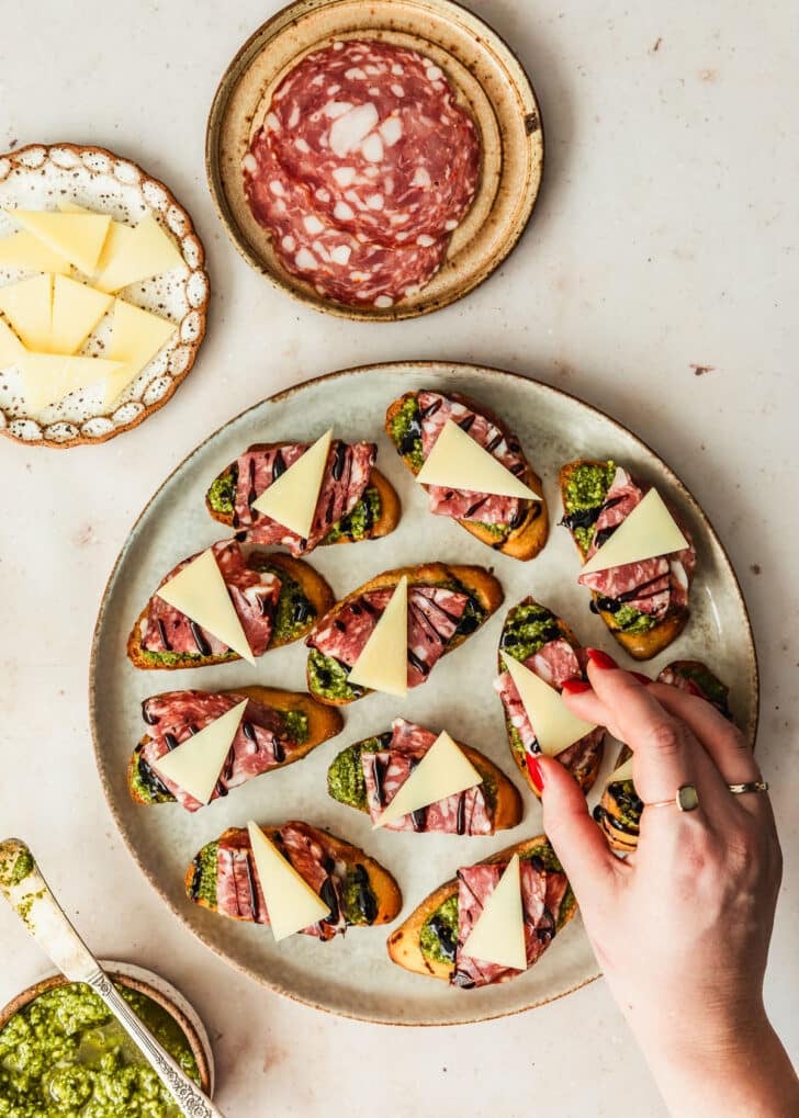 A hand placing slices of parmesan on a plate of soppressata pesto crostini next to brown bowls of pesto, soppressata, and parmesan with a tan marble background.