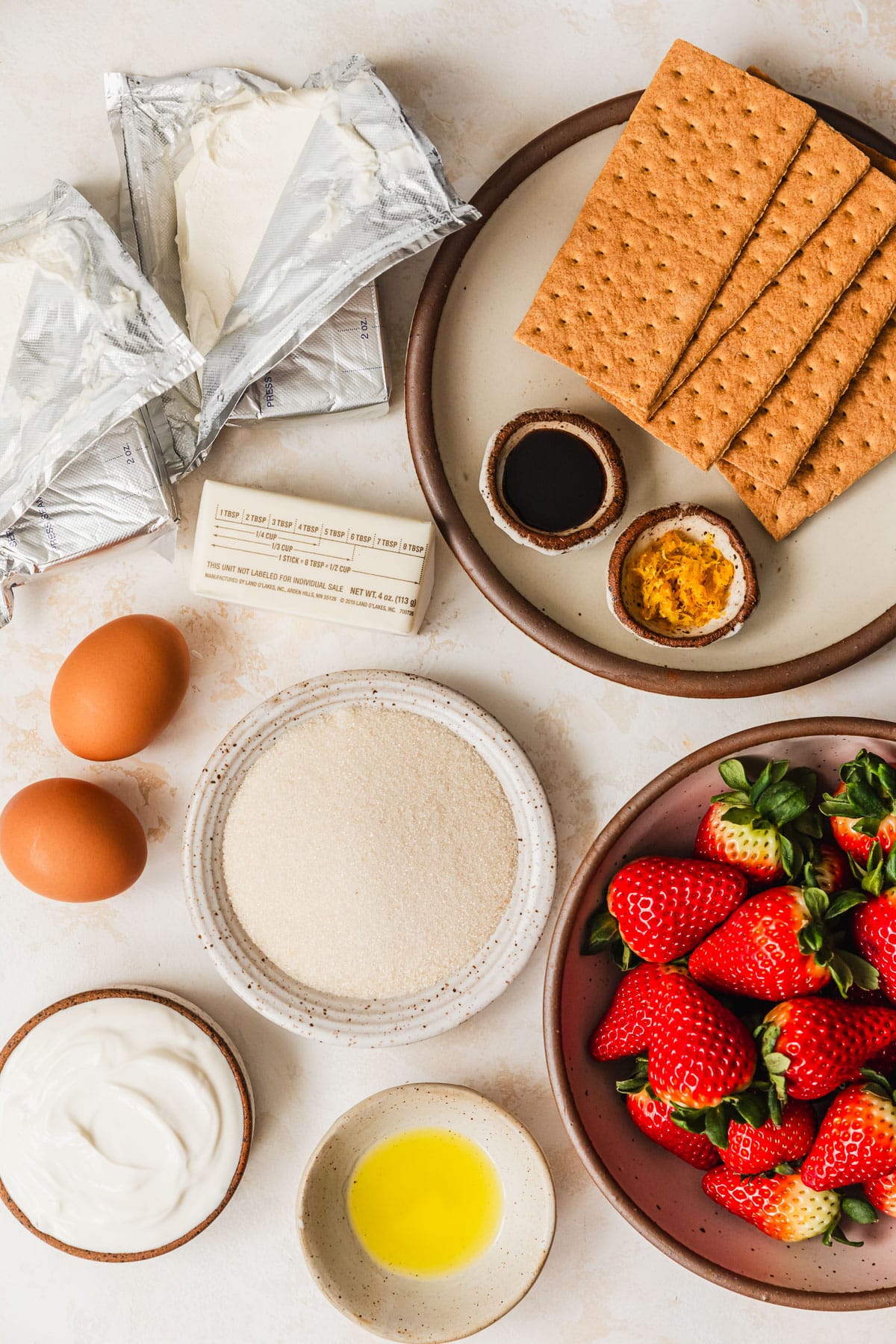 White and brown bowls of sugar, graham crackers, vanilla, lemon, strawberries, limoncello, and sour cream on a tan counter next to a stick of butter, eggs, and two blocks of cream cheese.