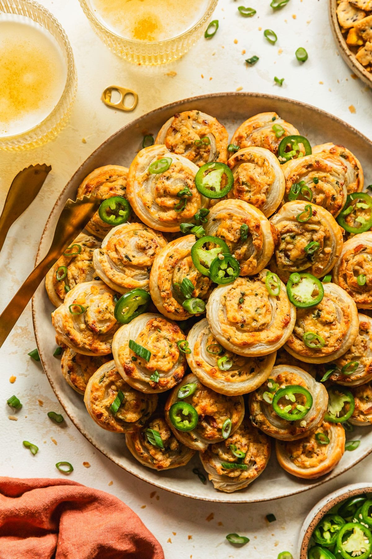 A beige plate of baked jalapeño popper pinwheels on a tan counter next to an orange linen, glasses of beer, and brown bowl of sliced jalapeños.