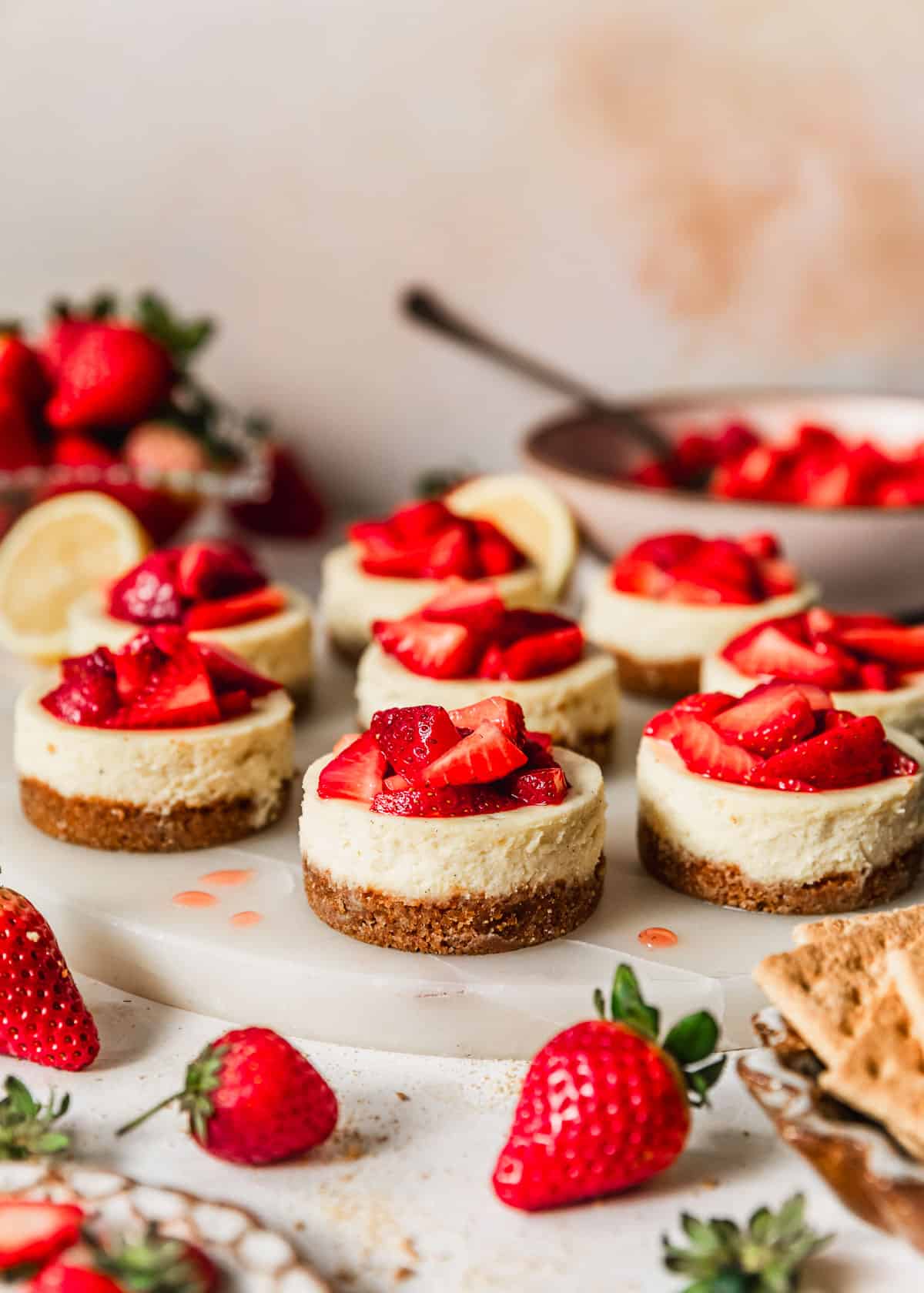 A marble tray of mini strawberry cheesecakes on a pink backdrop next to lemons, strawberries, and a pink bowl of berries.