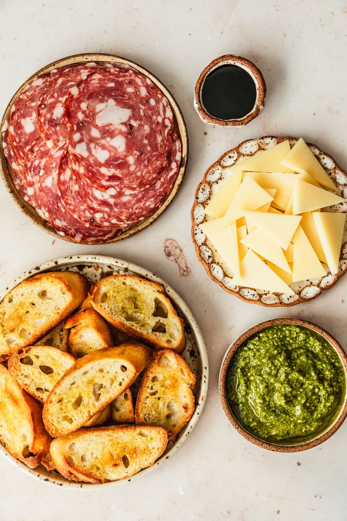 Brown and white bowls of soppressata, parmesan, pesto, crostini, and balsamic glaze on a tan counter.