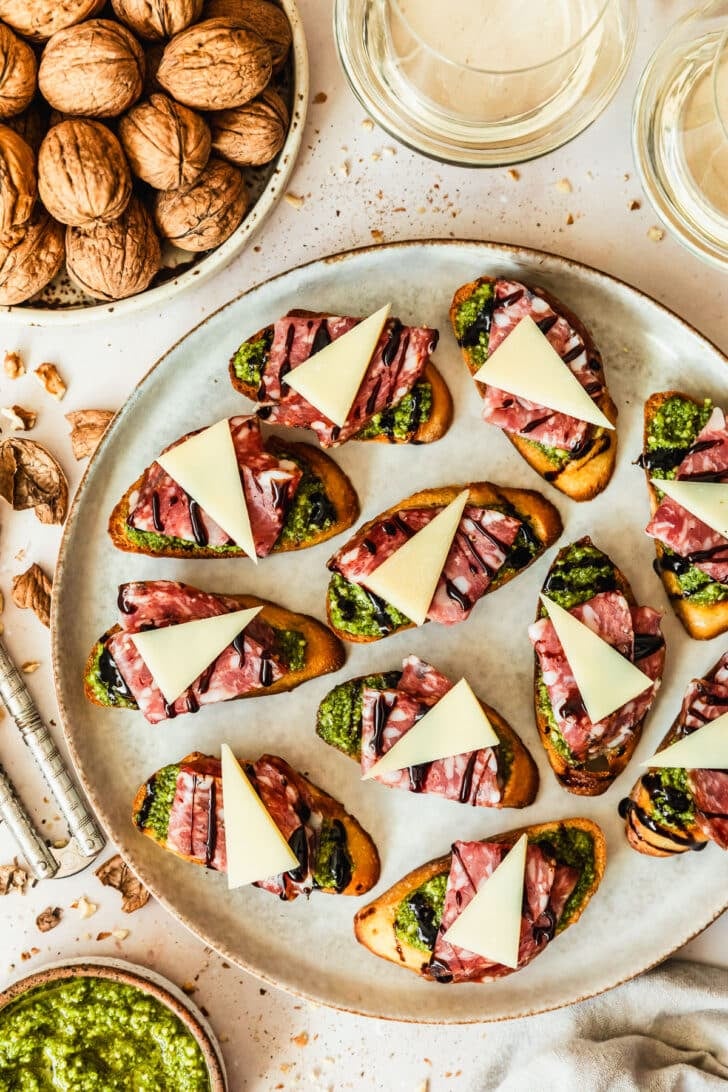 A grey platter of soppressata, parmesan, and pesto crostini on a tan counter next to glasses of white wine, a brown bowl of pesto, and a white bowl of walnuts.