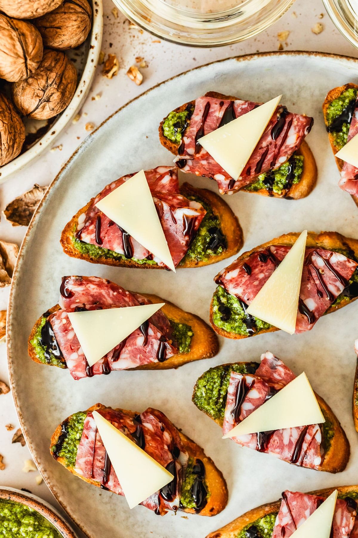 Soppressata, parmesan, and pesto crostini on a grey plate next to glasses of white wine, a white bowl of walnuts, and a brown bowl of pesto with a tan background.