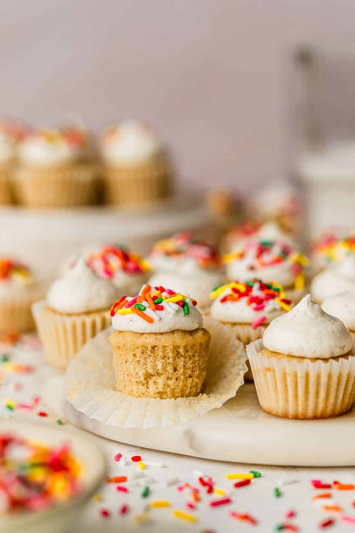 A white platter of mini vanilla cupcakes on a pink backdrop next to a white bowl of sprinkles and glass of milk.