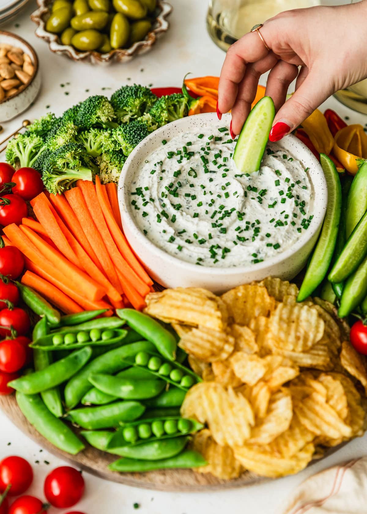 A hand dipping a cucumber slice in a white bowl of herb and garlic dip on a veggie tray next to glasses of white wine and white bowls of almonds and olives with a tan background.