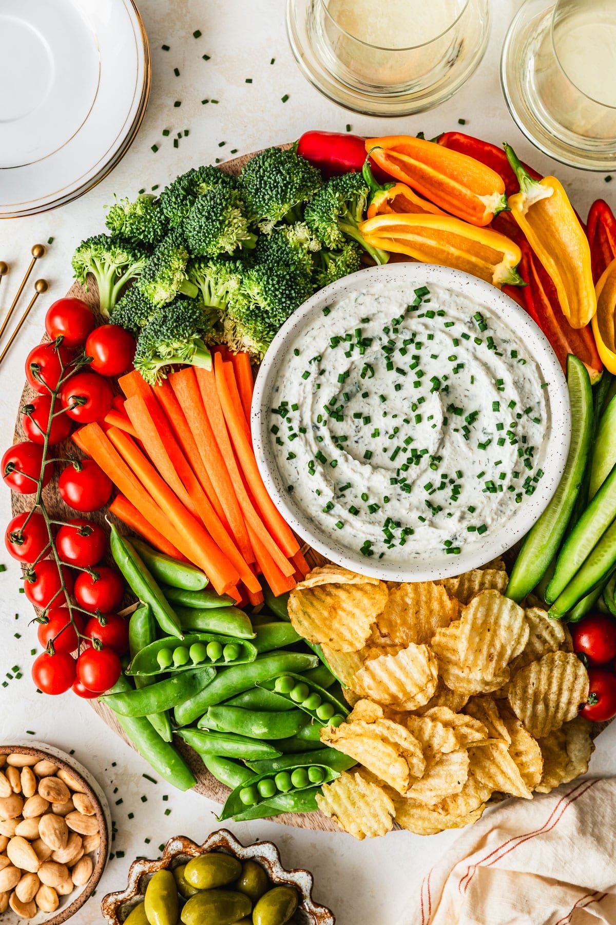 A white bowl of herb and garlic dip on a veggie platter next to glasses of white wine and white bowls of almonds and olives with a tan background.