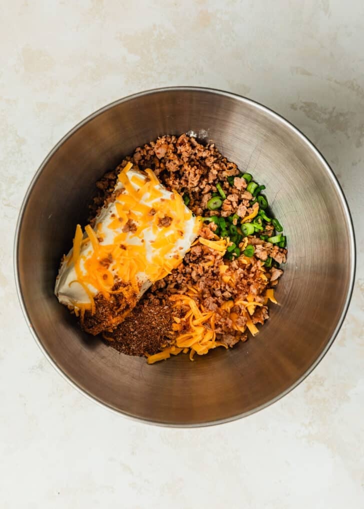 A silver bowl with cream cheese, sausage, cheese, green onions, and spices on a tan counter.