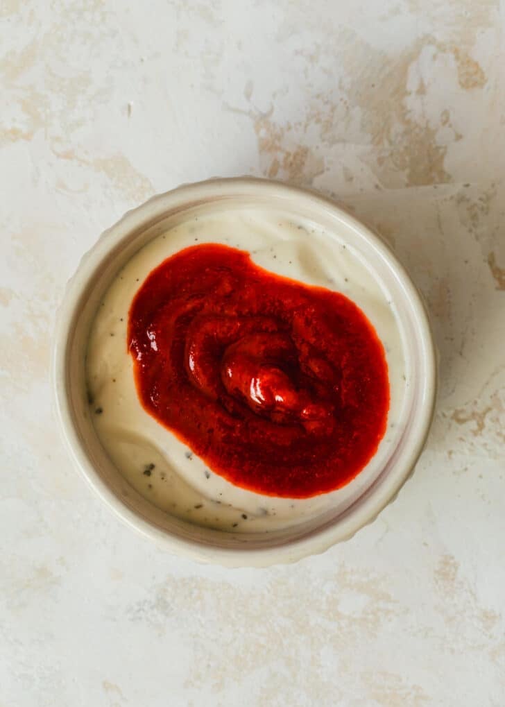A white bowl of ranch and Sriracha on a tan counter.