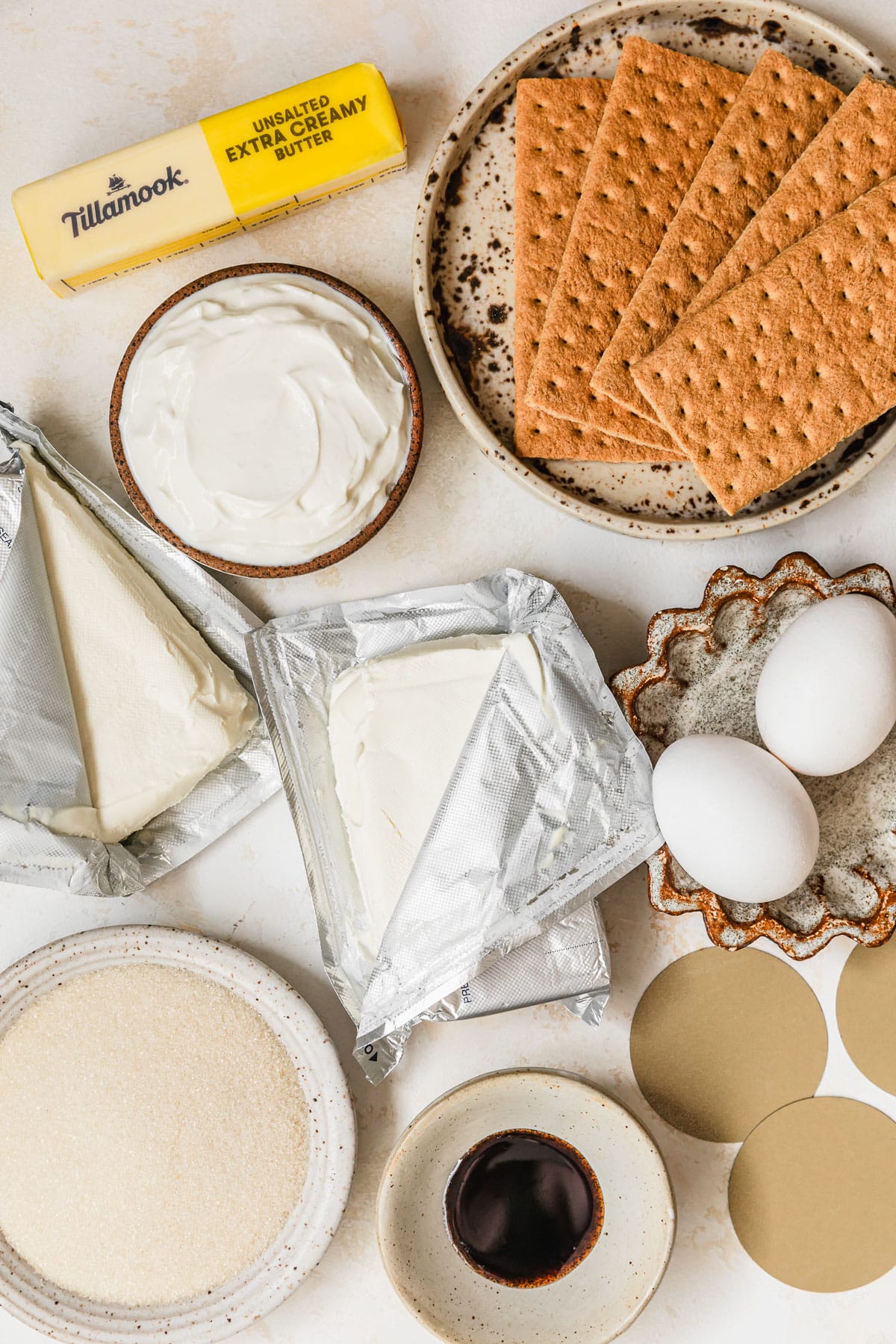 White and brown bowls of sour cream, graham crackers, cream cheese, eggs, sugar, and vanilla next to a stick of butter on a tan counter.