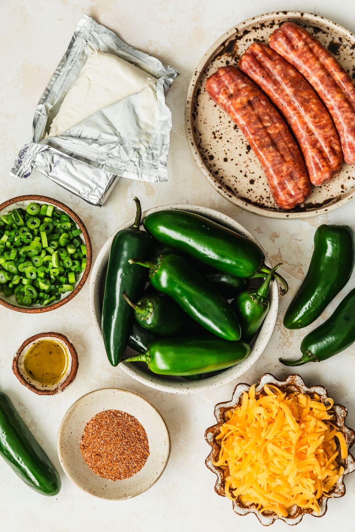 White and brown bowls of sausage, cream cheese, peppers, green onions, cheese, oil, and BBQ seasoning on a tan counter.