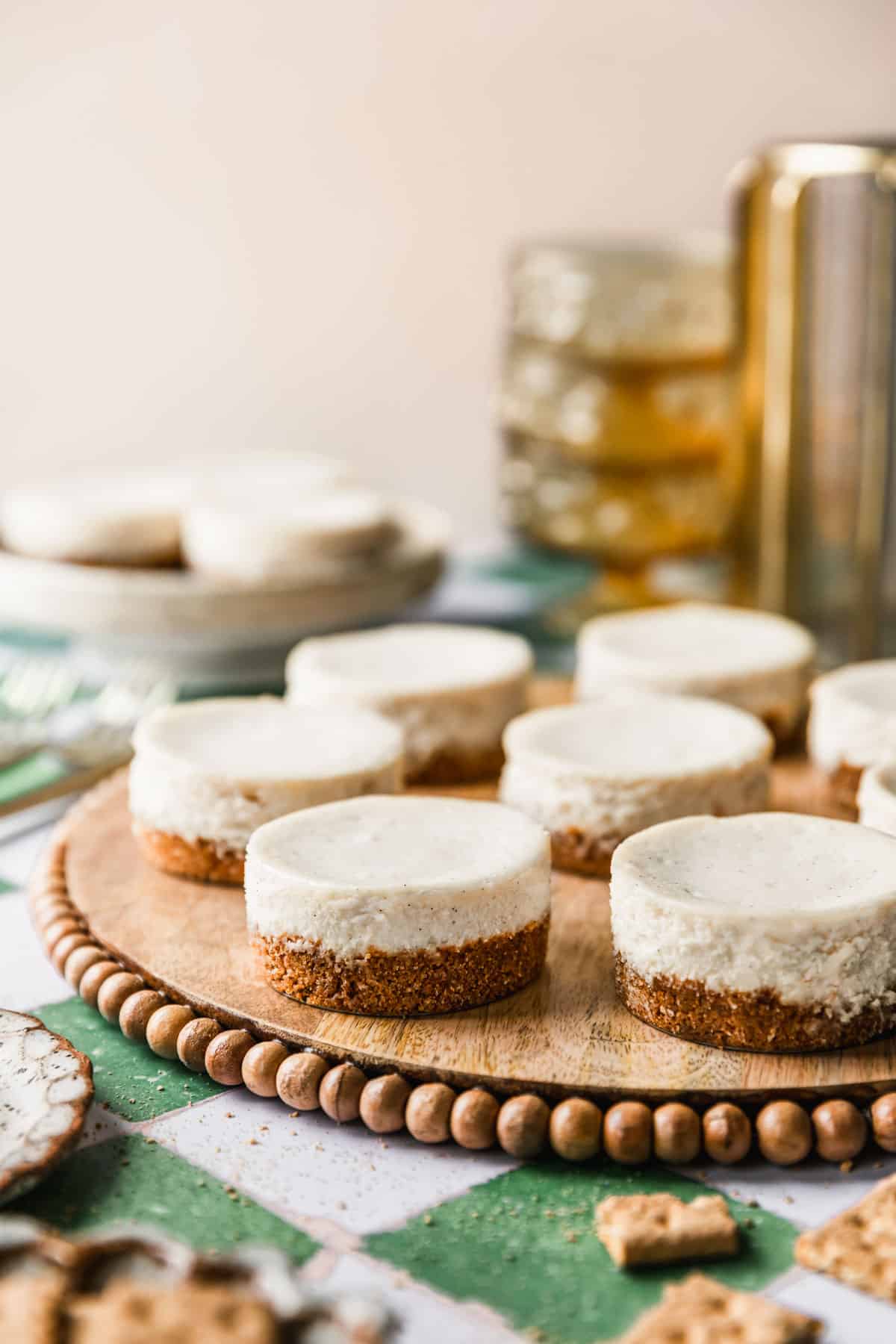 A wood tray of mini vanilla cheesecakes on a green and white tile backdrop next to glass yellow bowls, white plates, and graham crackers.