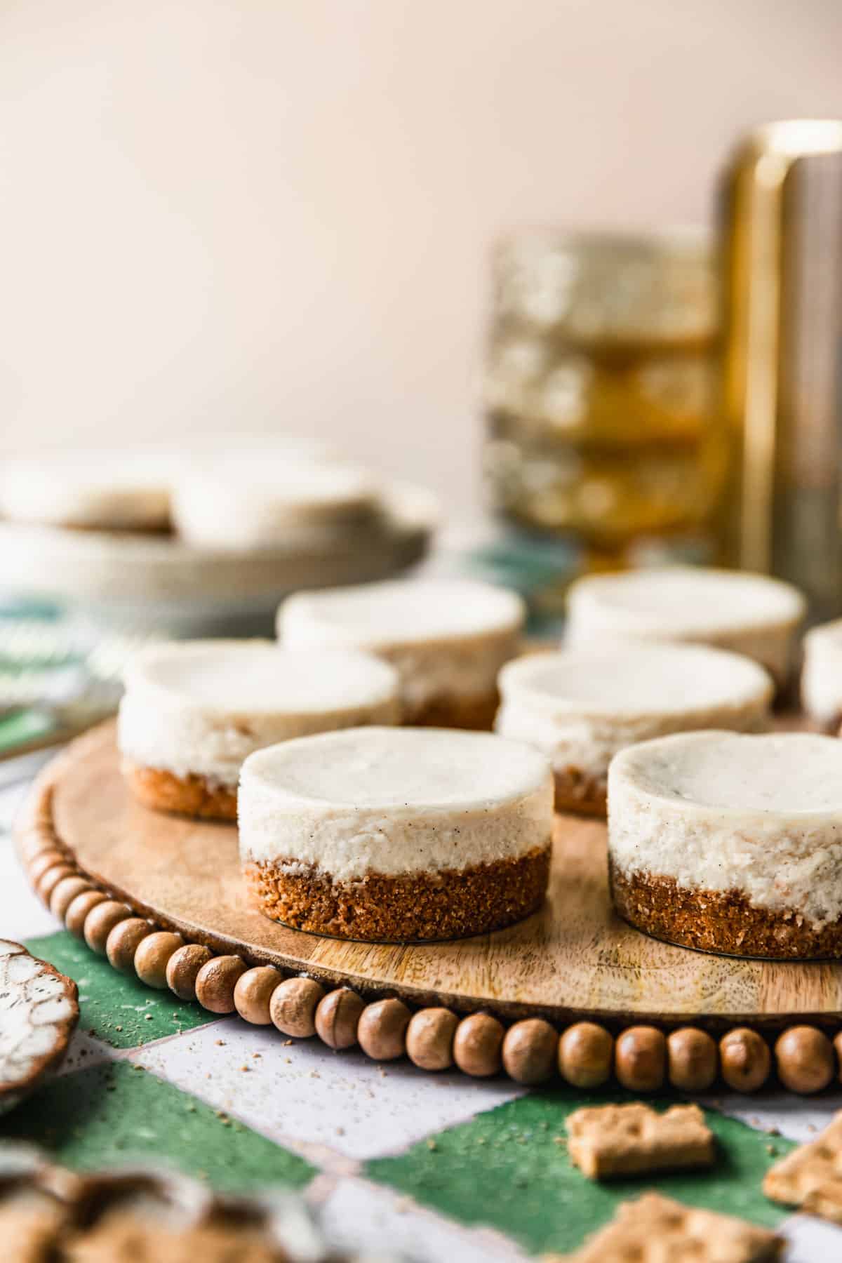 A wood tray of mini vanilla cheesecakes on a green and white tile counter next to graham crackers, yellow bowls, and a gold candle holder.