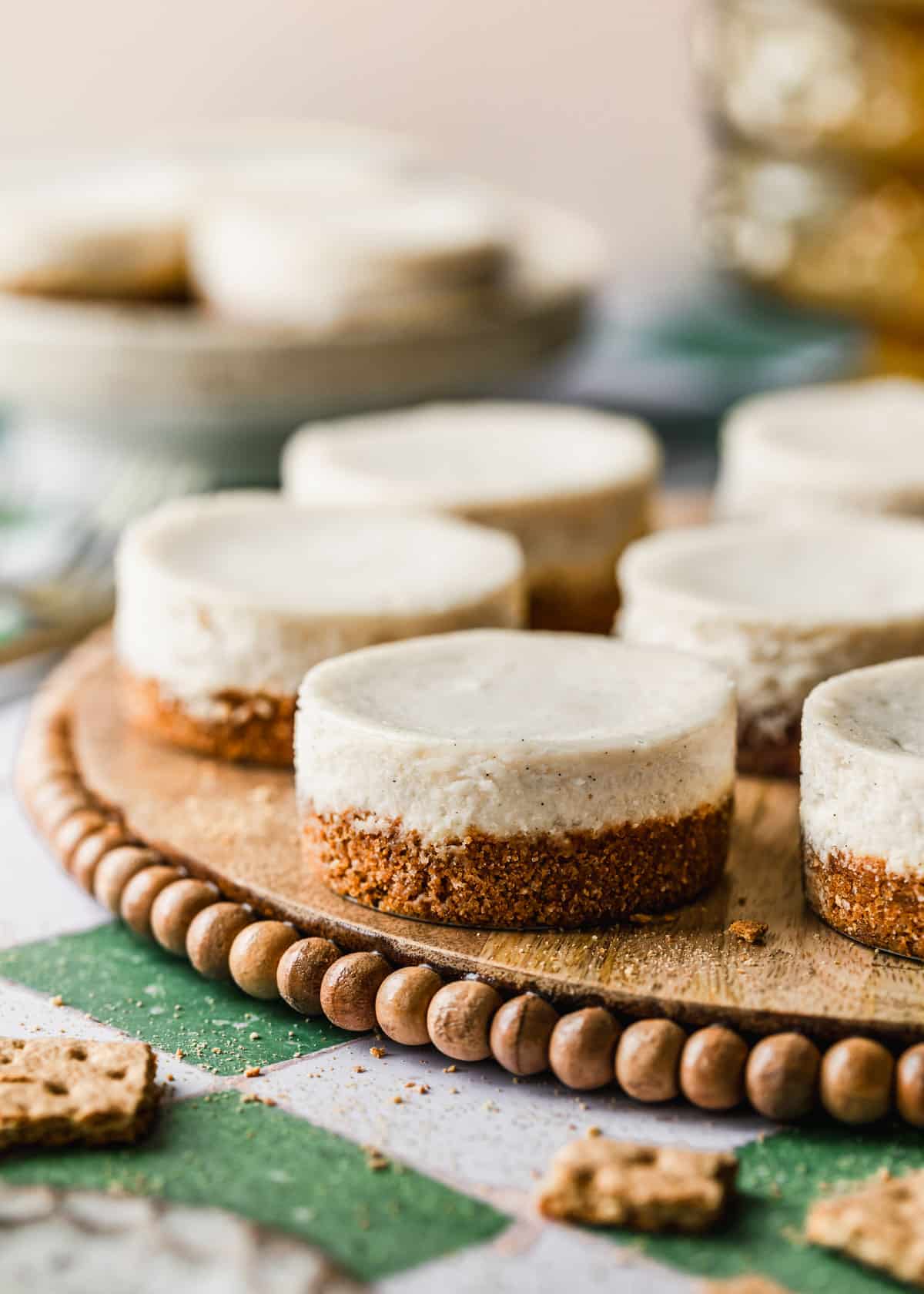 Mini vanilla cheesecakes on a wood tray next to graham crackers and yellow glass bowls with a green and white tile background.