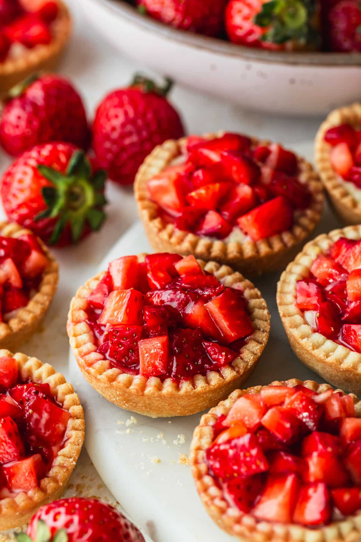 A white platter of mini strawberry custard tartlets next to a pink bowl of strawberries on a white background.