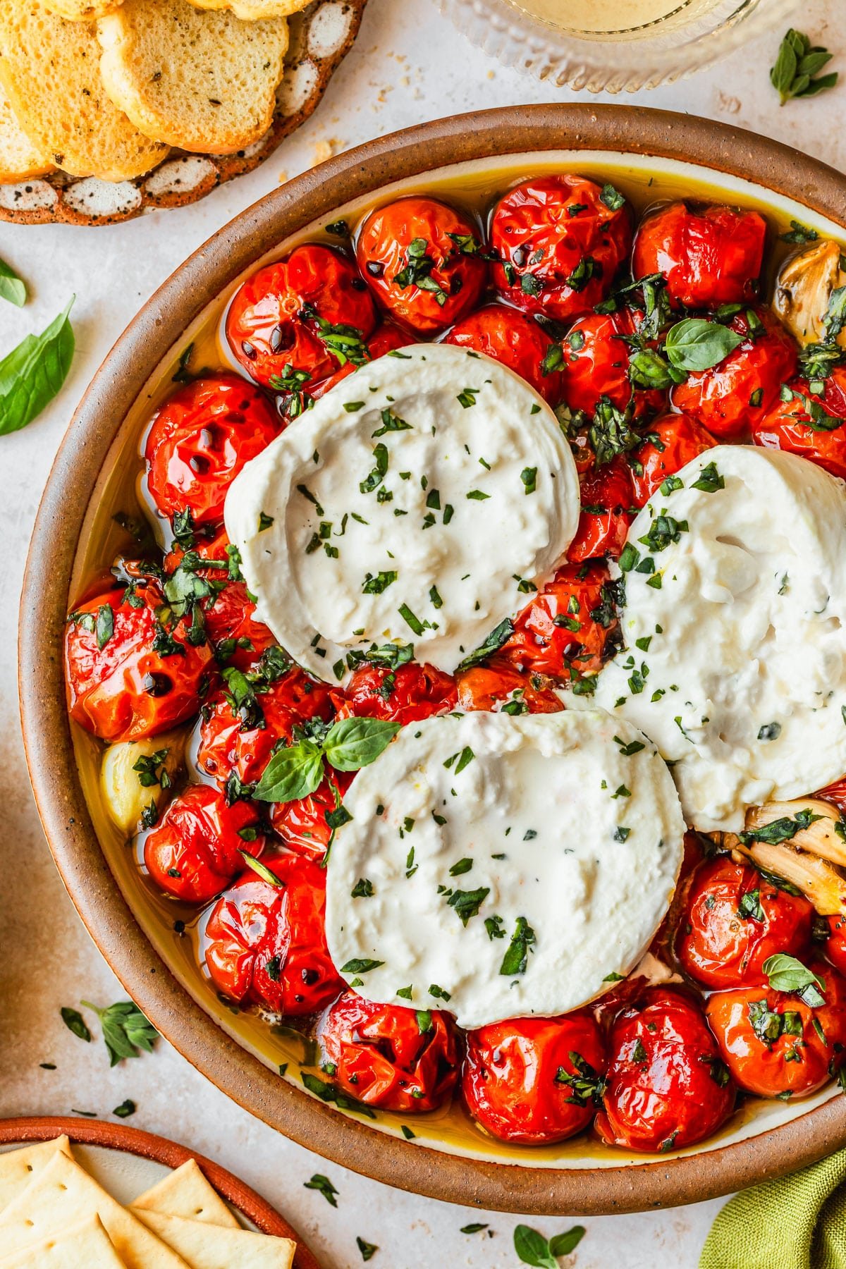 A brown platter of roasted tomato and burrata dip on a tan counter next to brown plates of crackers and glasses of white wine.