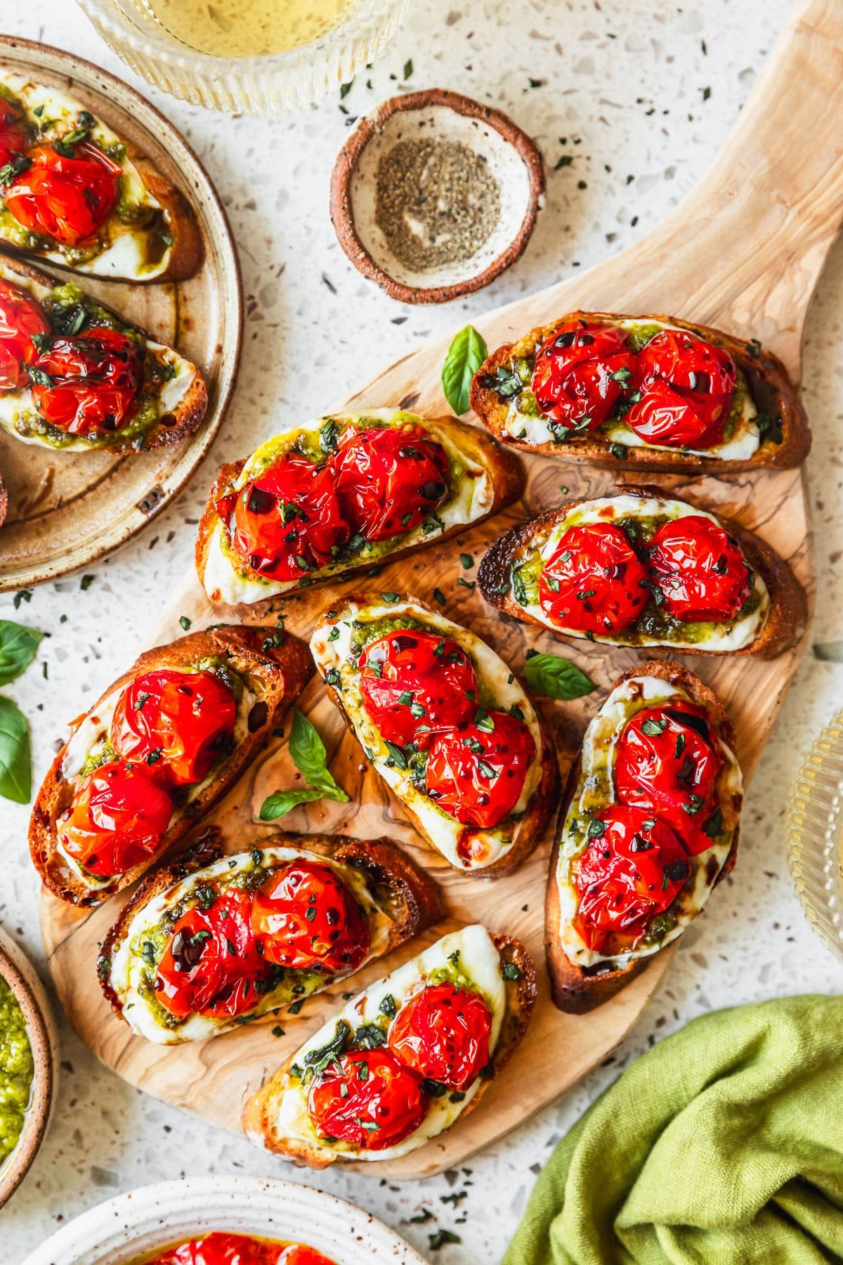 A wood board with baked Caprese crostini on a white terrazzo counter next to glasses of white wine, a green linen, and white bowls of tomatoes and pepper.