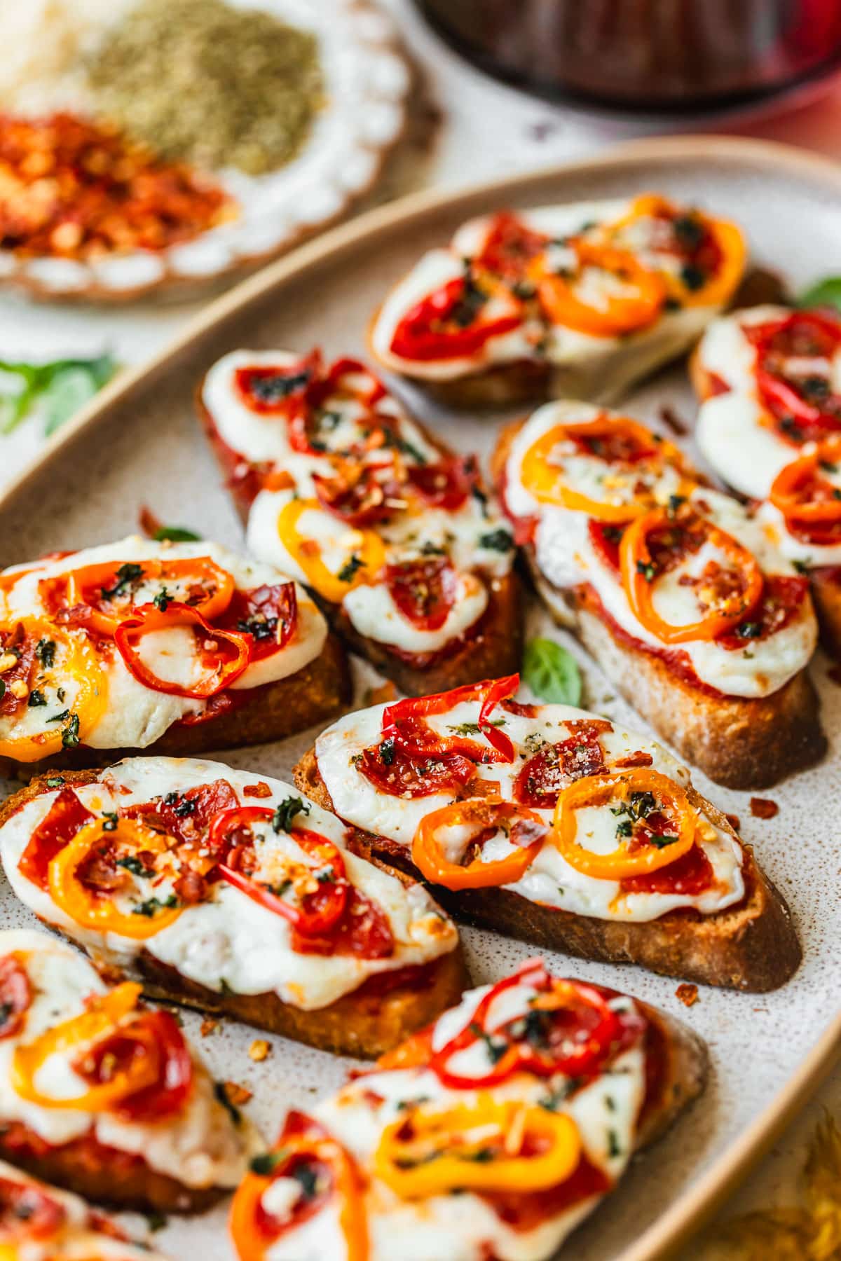 Rows of pepperoni pizza crostini on a stone platter next to glasses of red whine and a white plate of red pepper flakes and oregano.