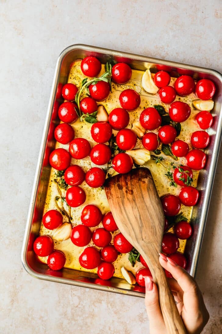 A hand using a spoon to mix tomatoes, garlic, and herbs on a sheet pan.