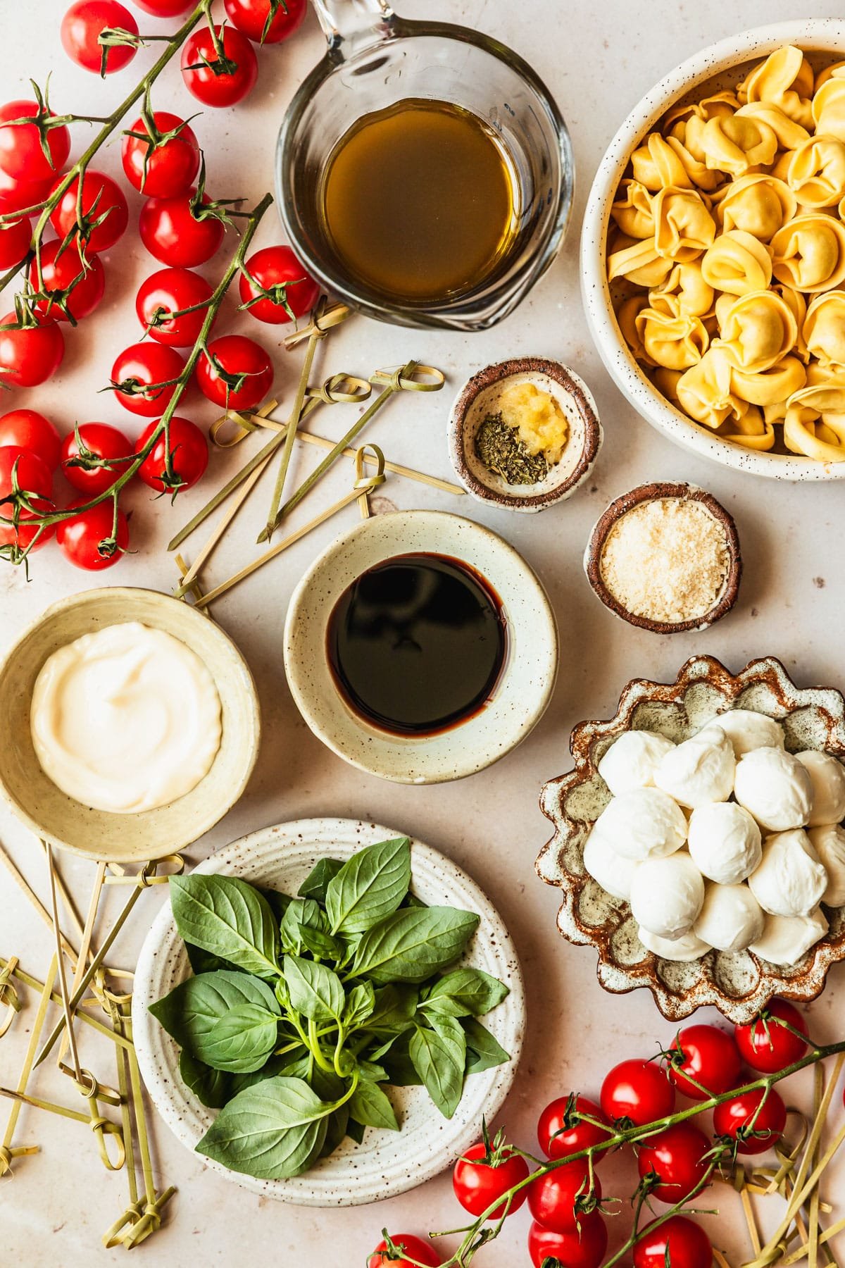 White and brown bowls of mozzarella, basil, balsamic, parmesan, mayo, tortellini, and herbs next to tomatoes on a tan marble counter.