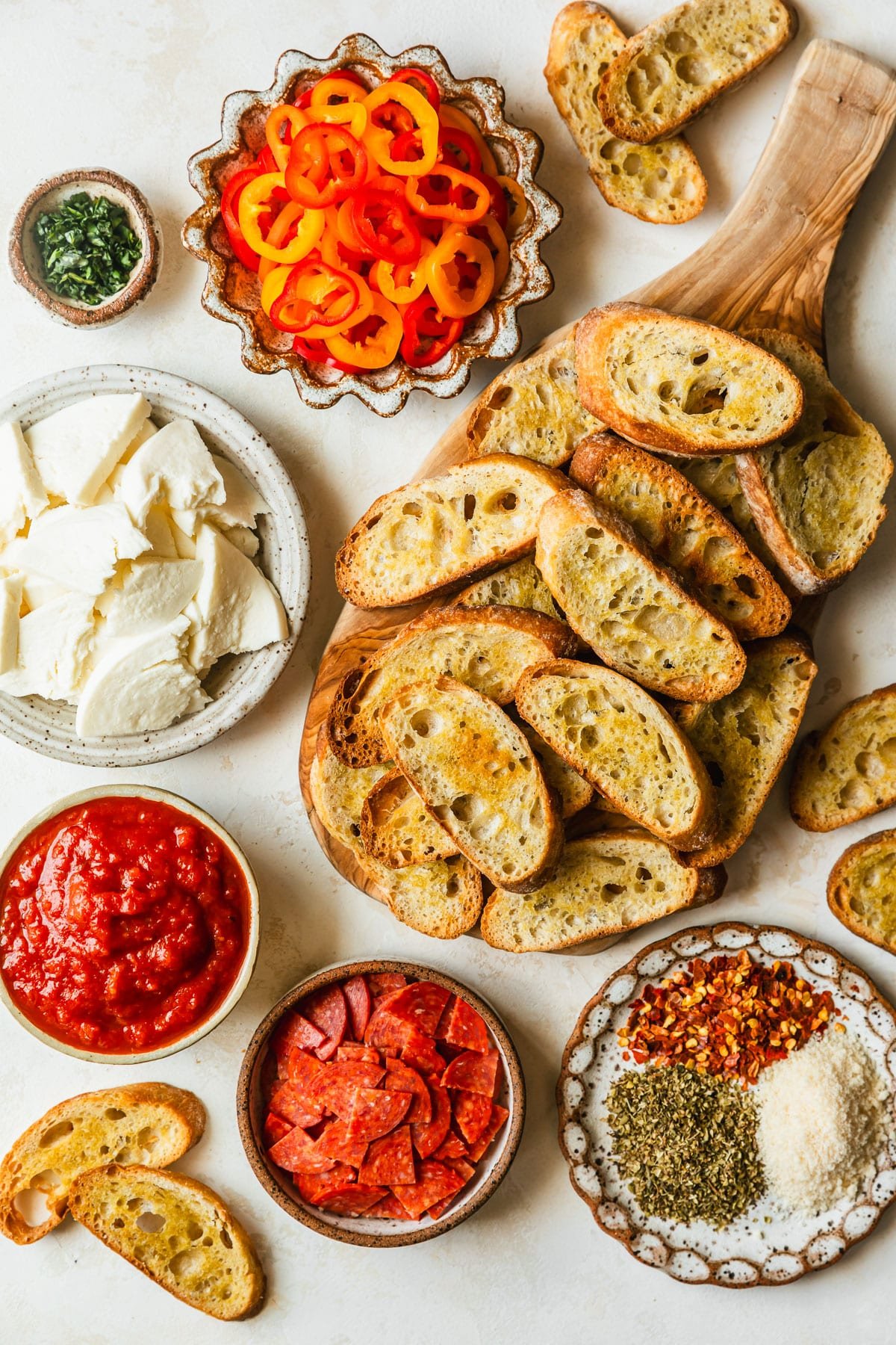Brown and white bowls of peppers, mozzarella, red sauce, pepperoni, and spices next to a wood board of toasts with a tan background.