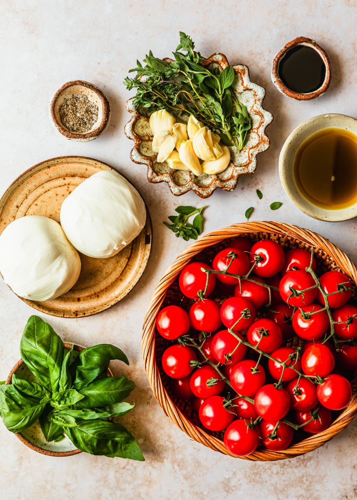 A basket of tomatoes next to brown and white bowls of balsamic, oil, herbs, garlic, pepper, burrata, and basil on a tan counter.
