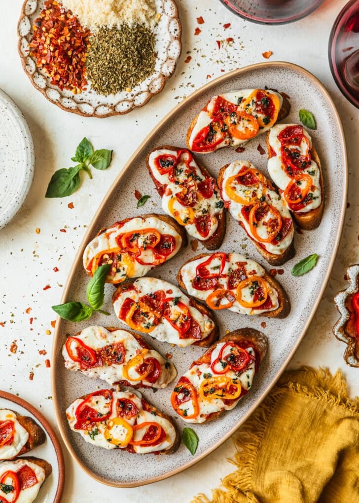 A stoneware platter of pepperoni pizza crostini next to glasses of red wine, a yellow linen, and a white plate of red pepper flakes, oregano, and parmesan with a white background.