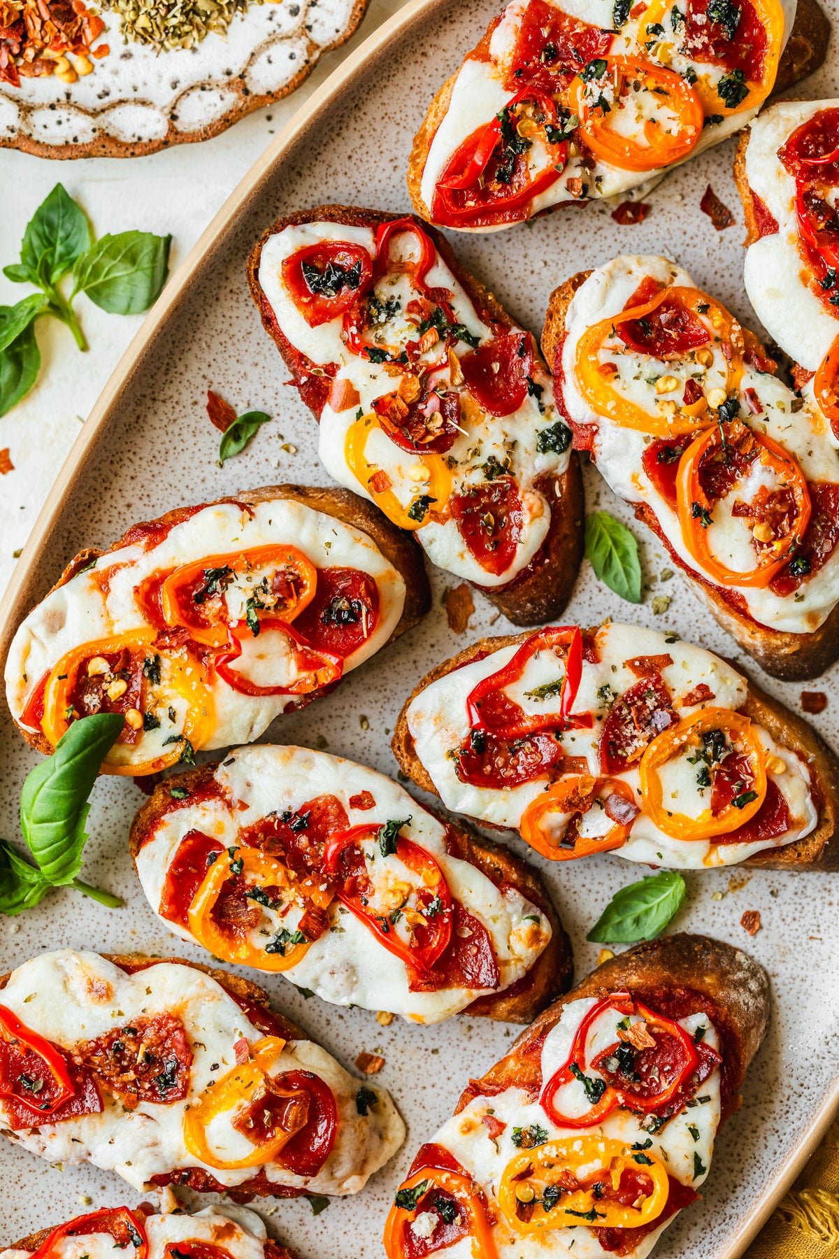 A stoneware platter of pizza crostini next to a white plate of crostini and fresh basil with a white background.