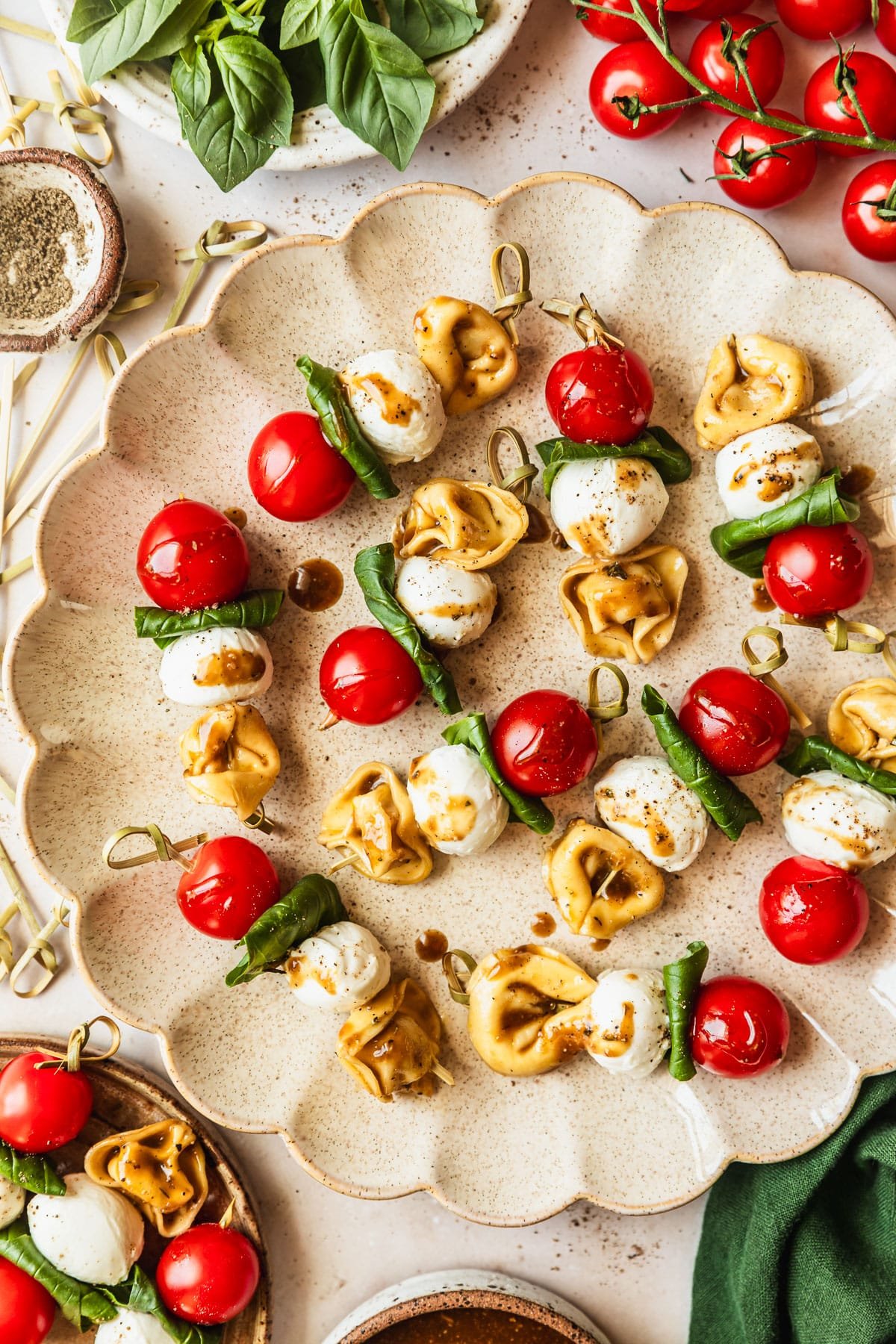 A tan platter of tortellini Caprese skewers on a marble counter next to a green linen and white bowls of balsamic dressing, basil, and pepper.