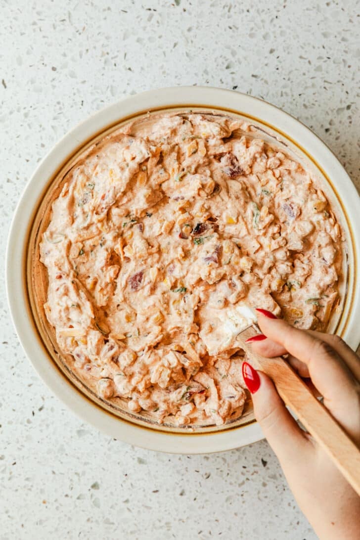 A hand using a rubber spatula to mix boat dip together in a white bowl.