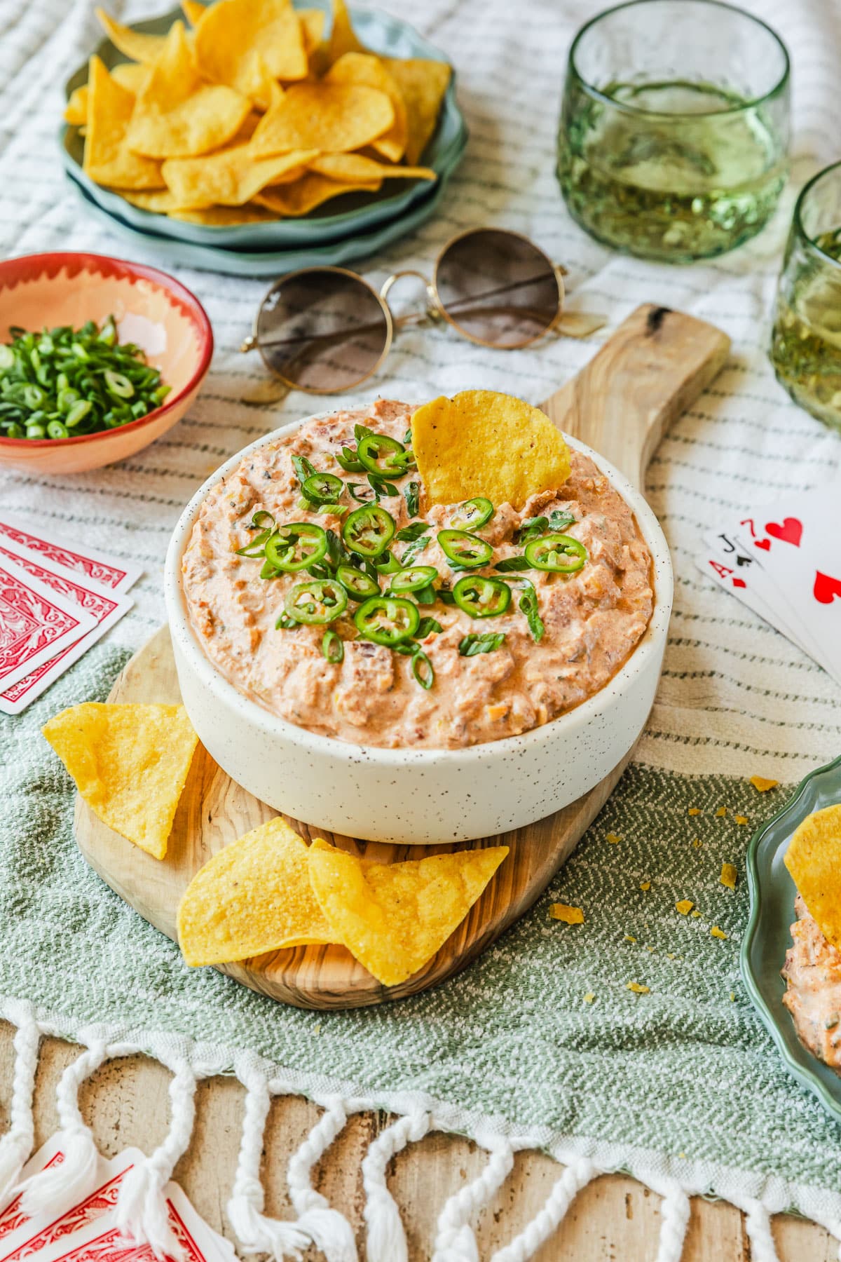 A white bowl of boat dip with corn on a wood board next to glasses of beer, a pink bowl of green onions, a blue bowl of corn chips, cards, and sunglasses with a white and green striped background.