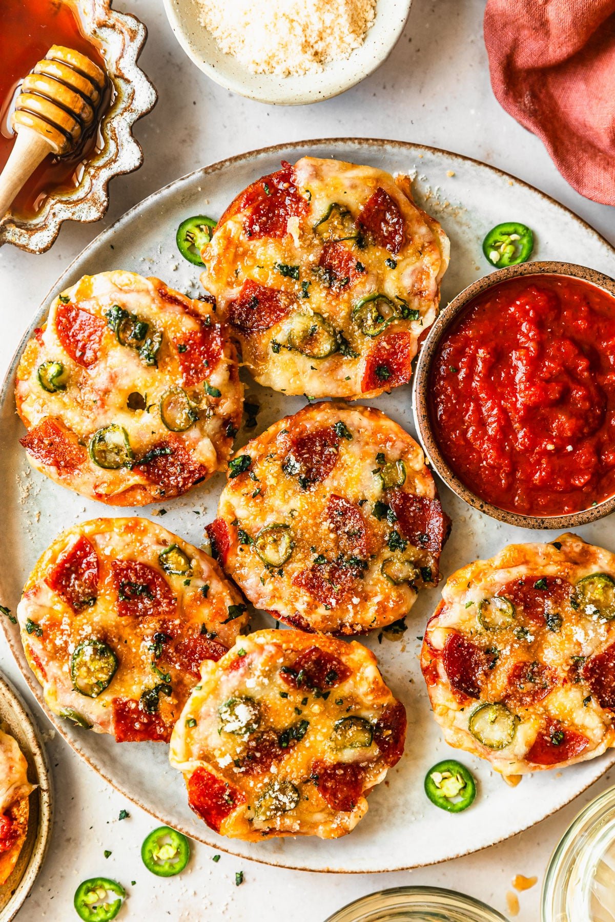 A grey platter of homemade pizza bagel bites on a tan counter next to white bowls of hot honey and parmesan, an orange linen, and glasses of white wine.