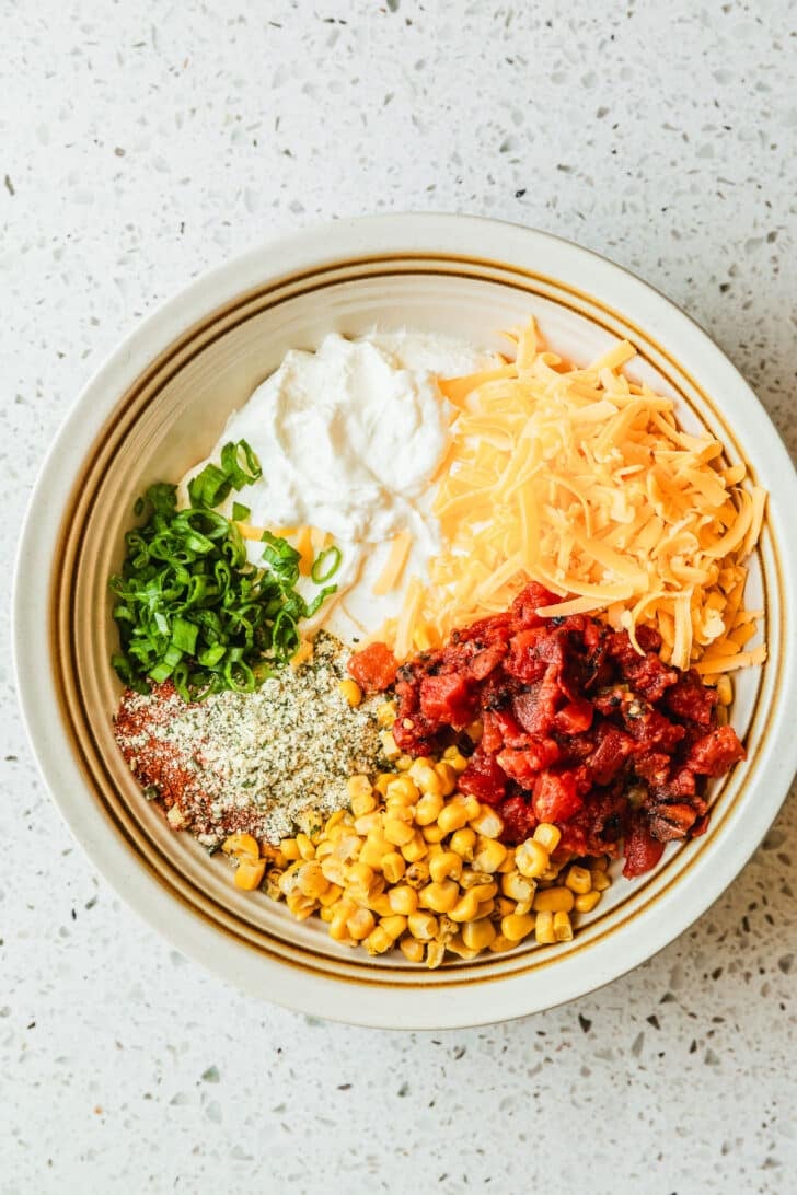 A white bowl of sour cream, seasonings, tomatoes, cheddar cheese, corn, and green onions on a white terrazzo counter.