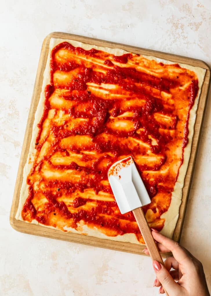 A hand using a rubber spatula to spread tomato sauce on puff pastry.
