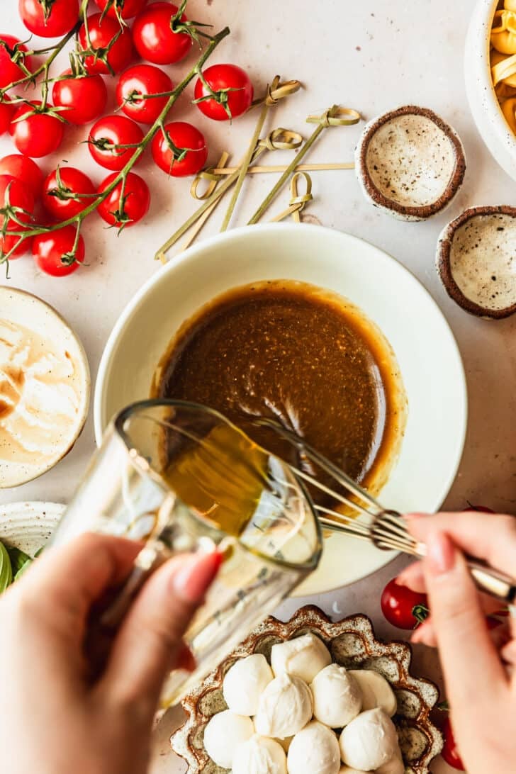 A hand pouring a glass measuring cup of olive oil into a white bowl of creamy balsamic dressing on a tan counter next to white bowls of mozzarella pearls, tortellini, and tomatoes.