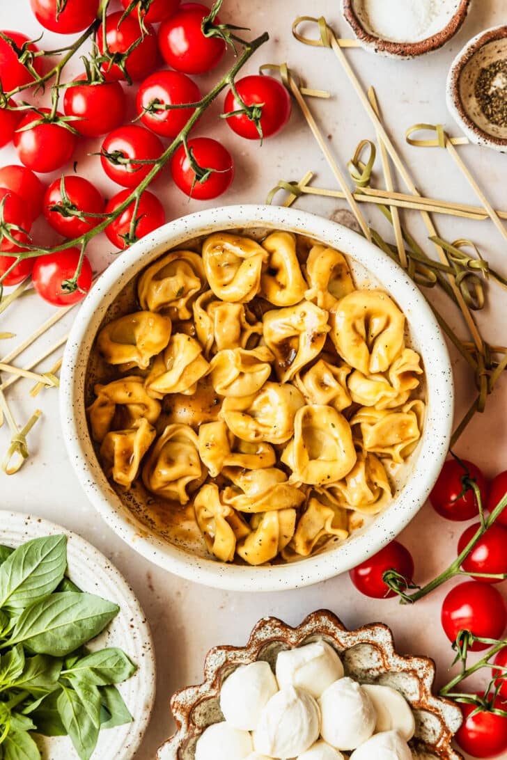 A white bowl of tortellini with balsamic dressing on a tan counter next to white bowls of mozzarella, basil, and tomatoes.