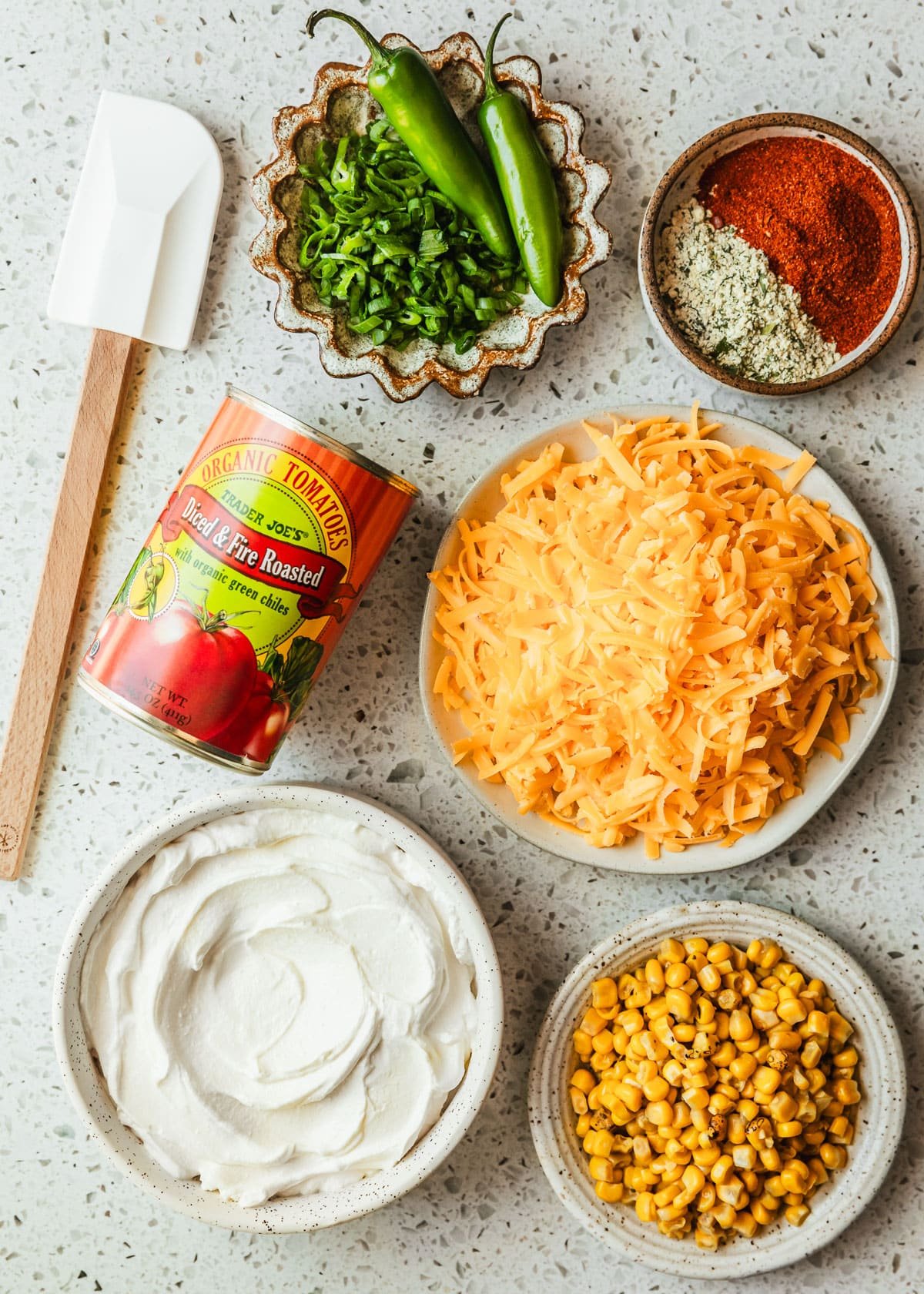 Brown and white bowls of cheddar cheese, sour cream, corn, ranch seasoning, taco seasoning, and green onions next to a can of tomatoes on a white terrazzo counter.